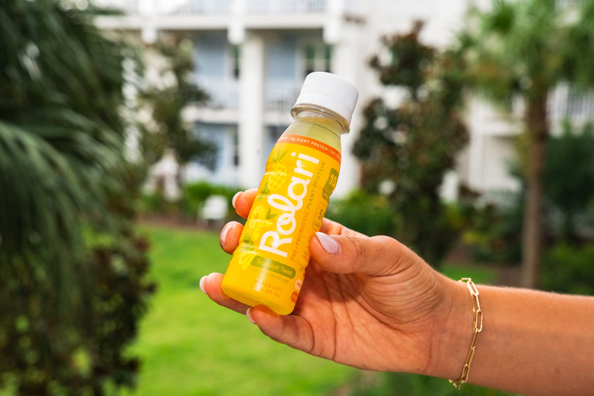 A hand holding a bottle of Rolari tropical twist against a blurred outdoor background of green grass and trees.