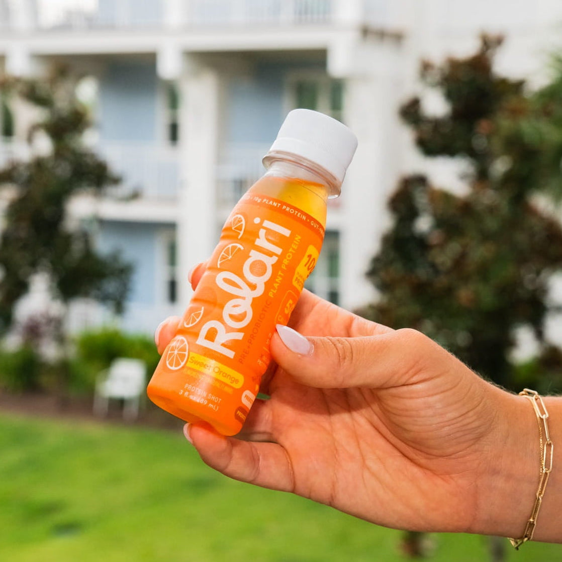 A hand holding an orange bottle of Rolari Orange Juice against a blurred outdoor background of a house and green lawn.