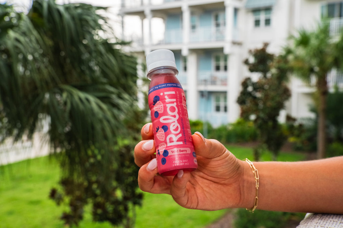 A hand holding a bottle of Rolari berry drink outdoors with palm trees and a building in the background.